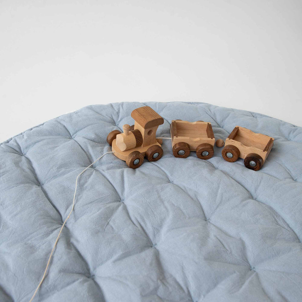 Angled view of the Sky Blue Cotton Quilted Play Mat with wooden toy trucks on top