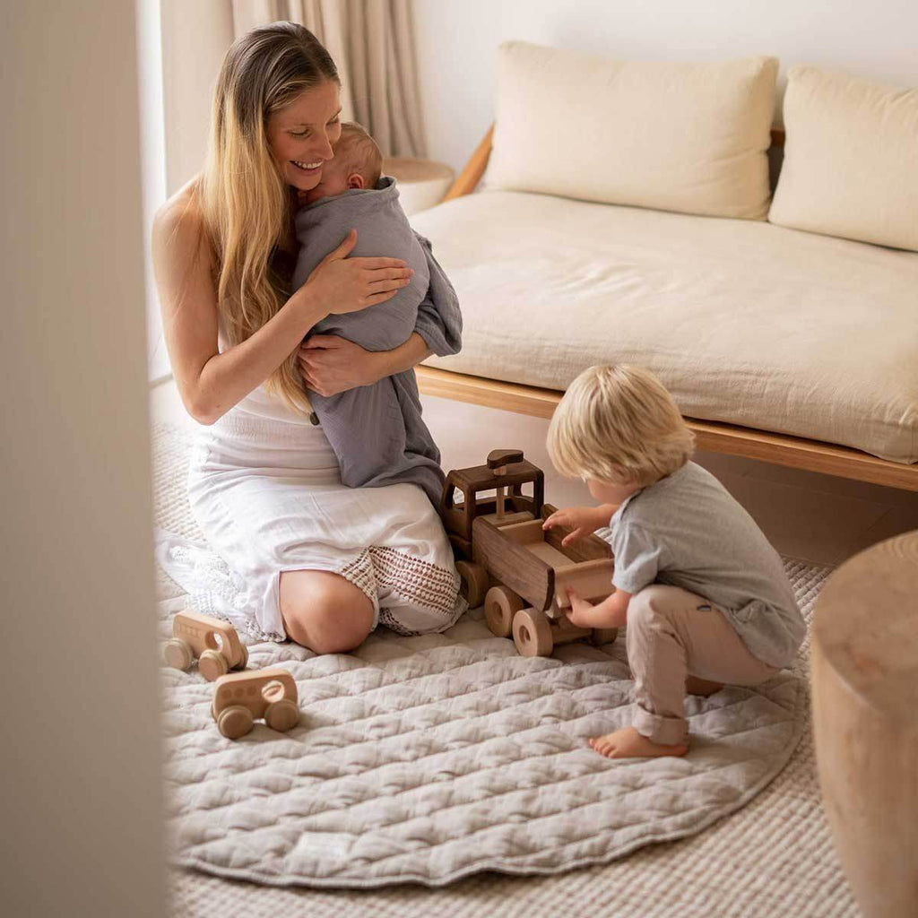 A toddler, mother and newborn baby playing with toys on the Natural Quilted Linen Play Mat